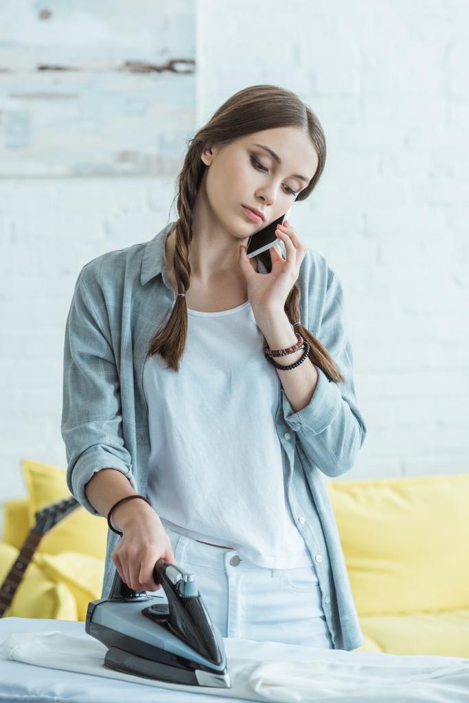 дівчина-підліток розмовляє на смартфоні під час прасування одягу
 - Фото, зображення