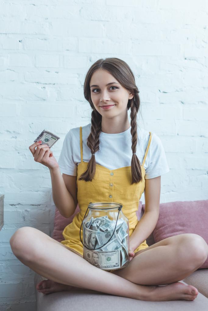 женщина-подросток кладет доллар банкноту в стеклянную банку для экономии, сидя на диване
 - Фото, изображение