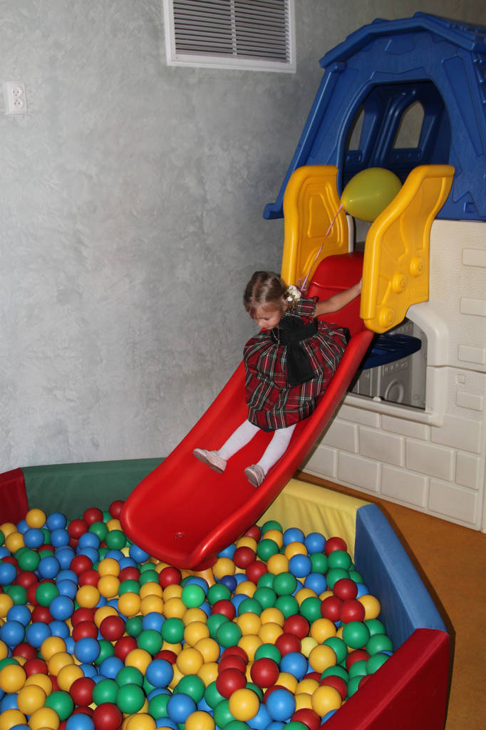 Маленькая девочка в клетчатом платье скатывается с детской горки в бассейн с мячами в детском саду
 - Фото, изображение