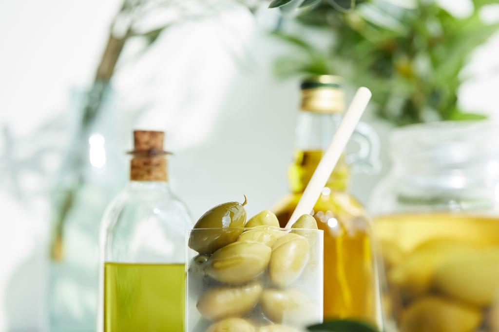 closeup shot of glass with spoon and green olives, jar, various bottles of aromatic olive oil with and branches on wooden tray - Photo, Image