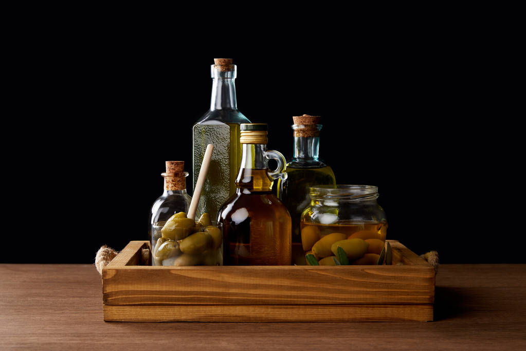 verschillende flessen van aromatische olijfolie en pot met groene olijven op houten tafel op zwarte achtergrond - Foto, afbeelding