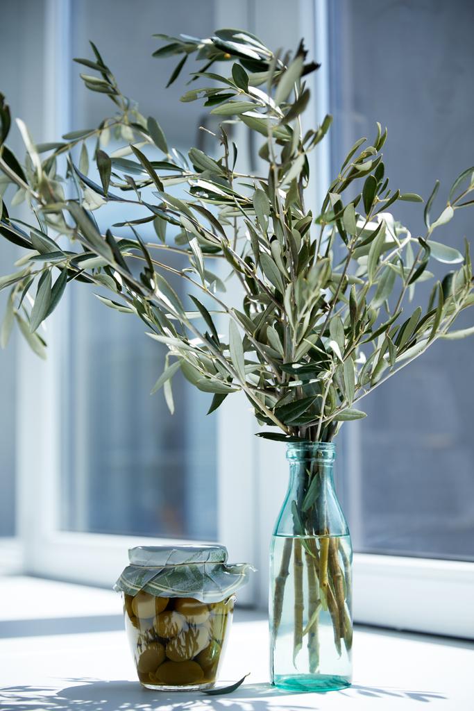 bottle with branches and jar with green olives on window sill - Photo, Image