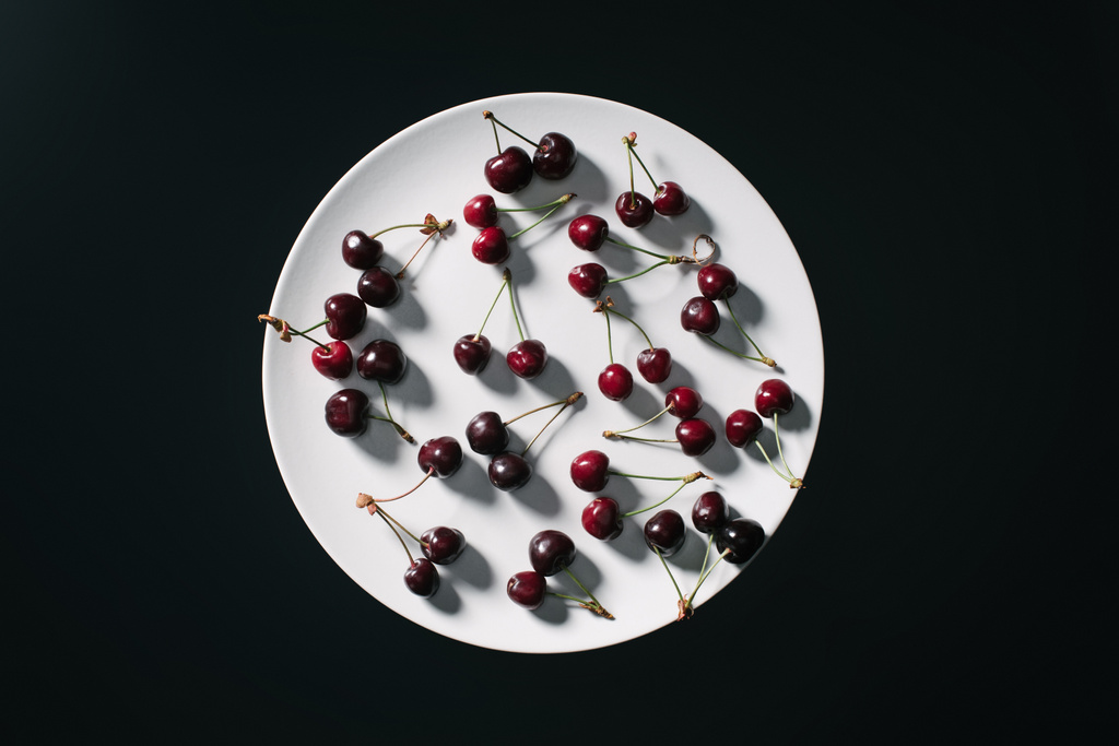 top view of fresh ripe red cherries on round white plate isolated on black - Photo, Image