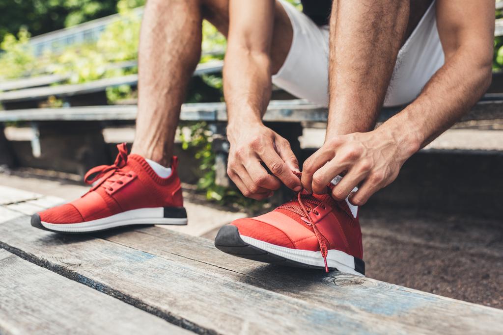 immagine ritagliata di sportivo allacciatura lacci delle scarpe mentre seduto sulla panchina al parco giochi sportivo
  - Foto, immagini