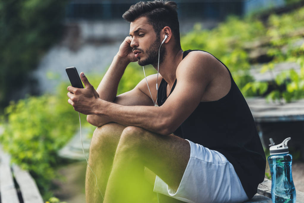 зосереджений спортсмен з пляшкою води, що слухає музику зі смартфонами на лавці на спортивному майданчику
 - Фото, зображення