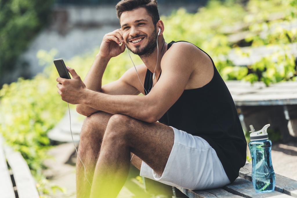 deportista sonriente con botella de agua escuchando música con smartphone y auriculares en el banco en el parque deportivo
 - Foto, Imagen