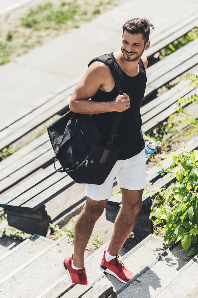 deportista sonriente va arriba con botella de agua y bolsa para equipos deportivos en el patio de recreo deportivo
  - Foto, Imagen