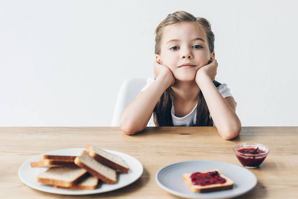 petite écolière réfléchie avec des toasts et de la confiture pour le petit déjeuner en regardant la caméra isolée sur blanc
 - Photo, image