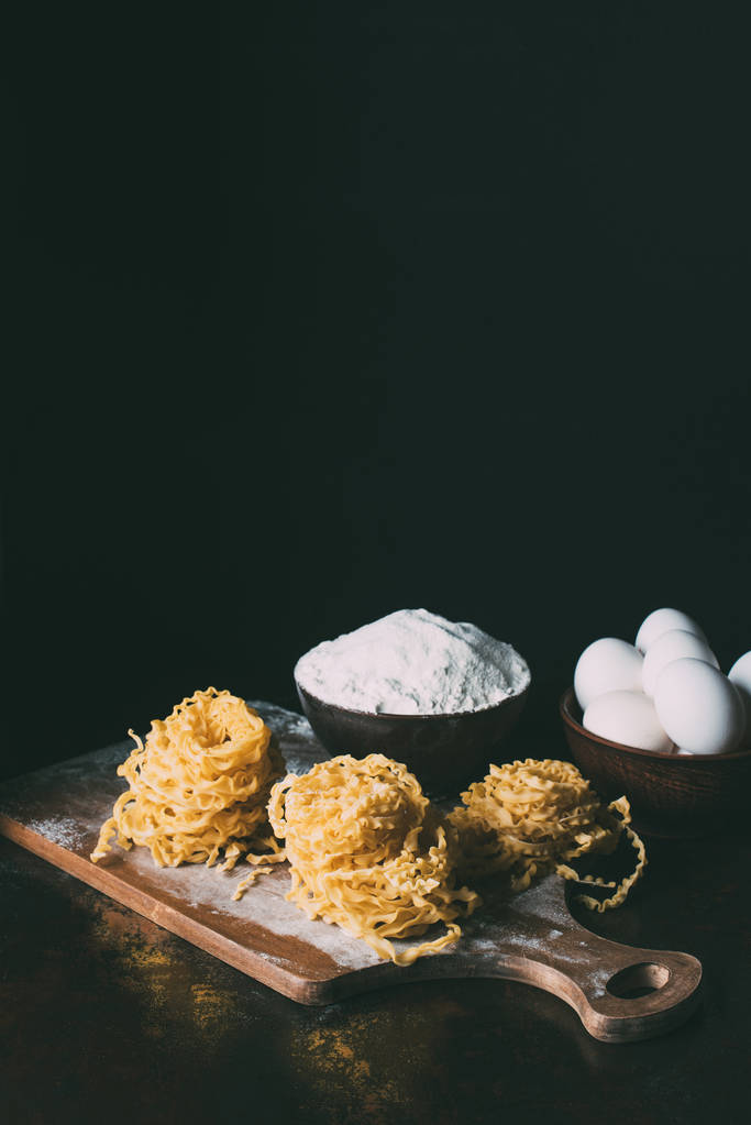 vista frontal de la tabla de cortar con pasta cruda, cuencos con harina y huevos en la mesa sobre fondo negro
  - Foto, Imagen
