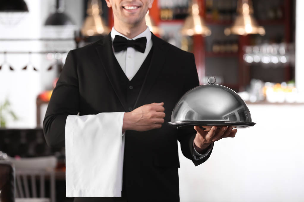 Официант в элегантной униформе держит металлический поднос и клош на рабочем месте
 - Фото, изображение