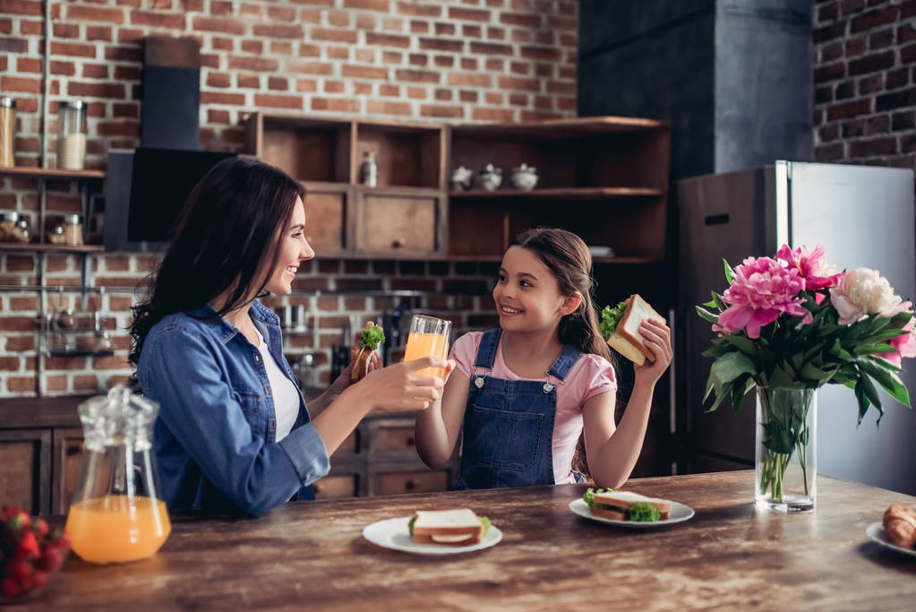 family of mother and daughter eating sandwiches with orange juice for breakfast in the kitchen - Photo, Image