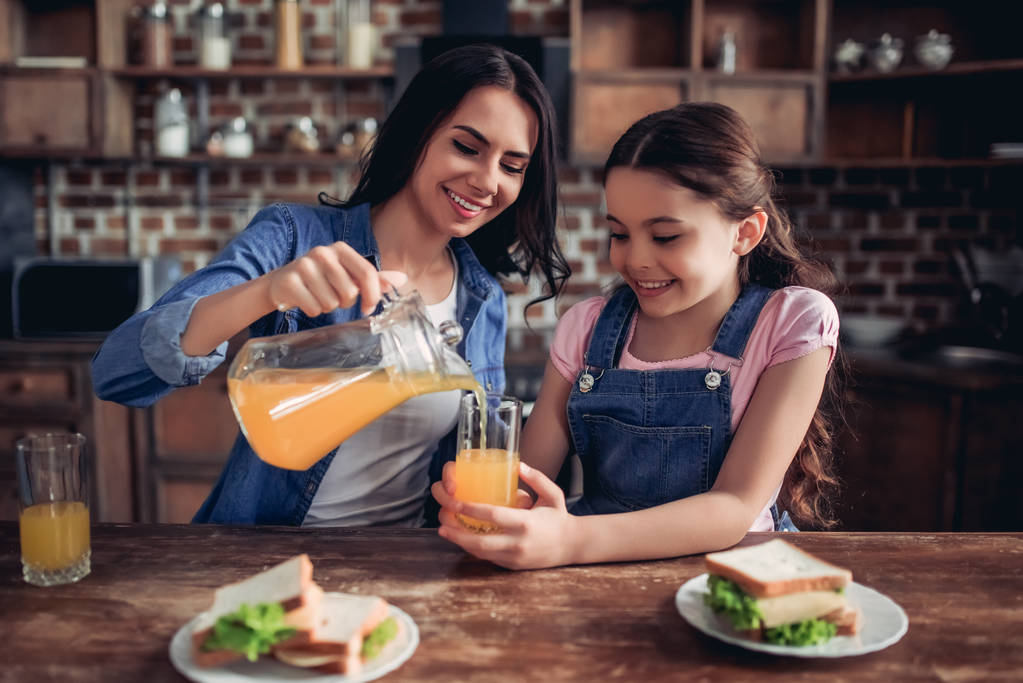 портрет улыбающейся матери, наливающей апельсиновый сок в стакан для дочери на кухне
 - Фото, изображение