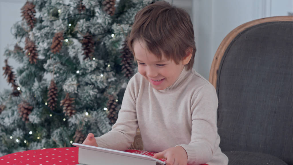 Uśmiechnięty chłopiec dziecko siedzi na krześle i grając z tabletu w czasie Bożego Narodzenia - Zdjęcie, obraz