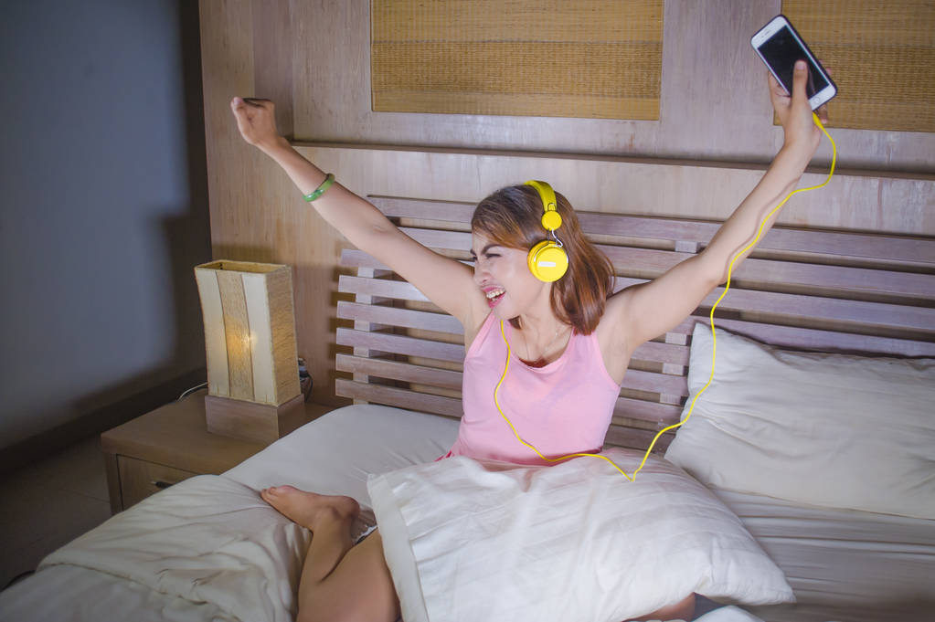 携帯電話のダンスと歌クレイジー ライフ スタイル コンセプトで彼女の寝室の家で興奮してインターネットで音楽を聞いて黄色のヘッドフォンが付いているベッドの魅力的で幸せな少女 - 写真・画像
