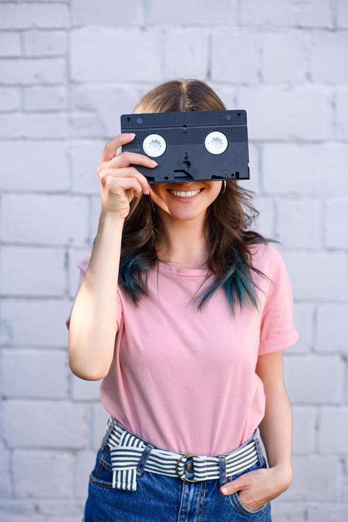 vista oscura de la mujer sonriente cubriendo los ojos con retro video cassette en la mano contra la pared de ladrillo blanco
 - Foto, imagen