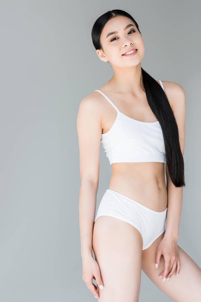 glimlachend aantrekkelijke Aziatische vrouw in witte lingerie poseren op een grijze achtergrond  - Foto, afbeelding