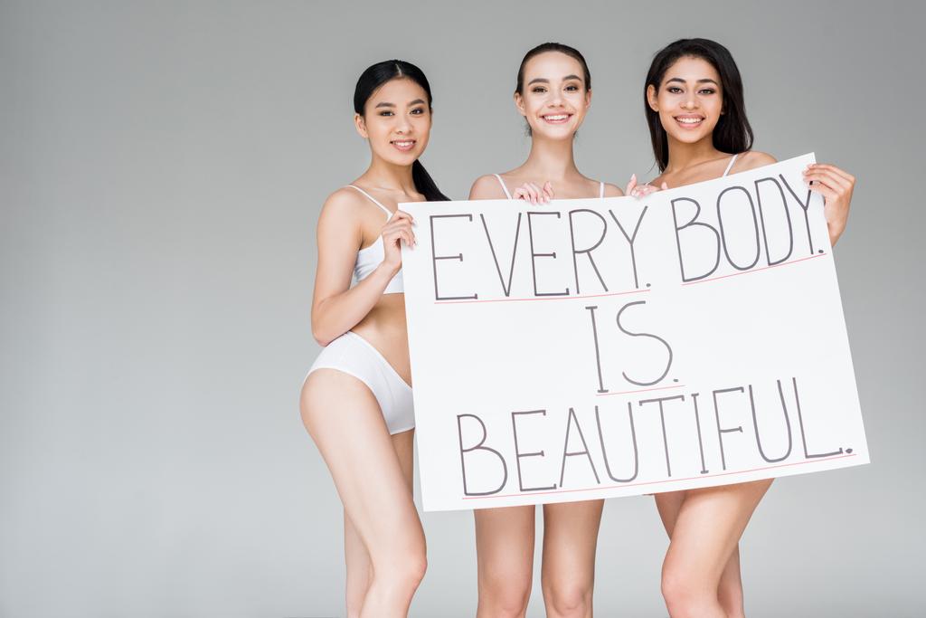 uśmiechający się wielokulturowym kobiet trzymając transparent z napisem każde ciało jest piękne na białym tle na szarym tle  - Zdjęcie, obraz
