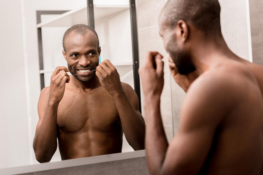 šťastný mladý muž čištění zubů dentální nit při pohledu na zrcadlo v koupelně - Fotografie, Obrázek
