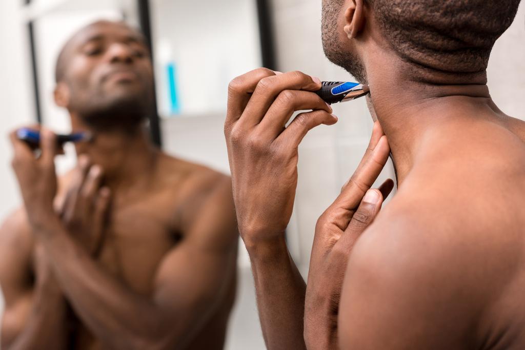 africano americano giovane barba da barba con rasoio elettrico mentre si guarda specchio in bagno
 - Foto, immagini