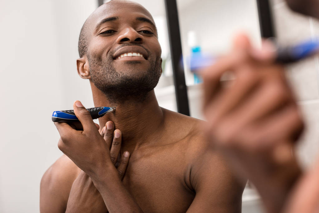 uśmiechający się młody człowiek golenia brody z golarka elektryczna, patrząc w lustro w łazience - Zdjęcie, obraz