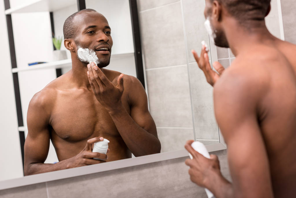 atractivo joven aplicando gel de afeitar mientras mira el espejo en el baño
 - Foto, imagen