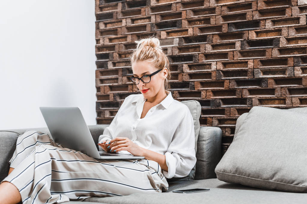 Beautiful blonde serious Caucasian woman sitting on couch at home and typing on laptop. - Photo, Image