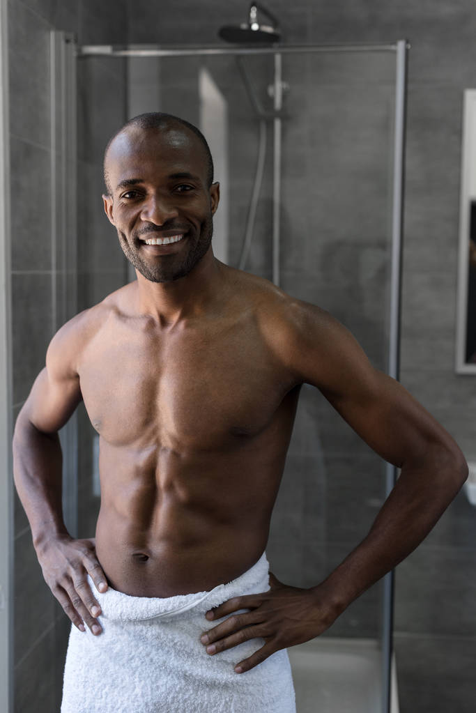 bel homme afro-américain à poitrine nue en serviette debout avec les mains sur la taille et souriant à la caméra dans la salle de bain
 - Photo, image