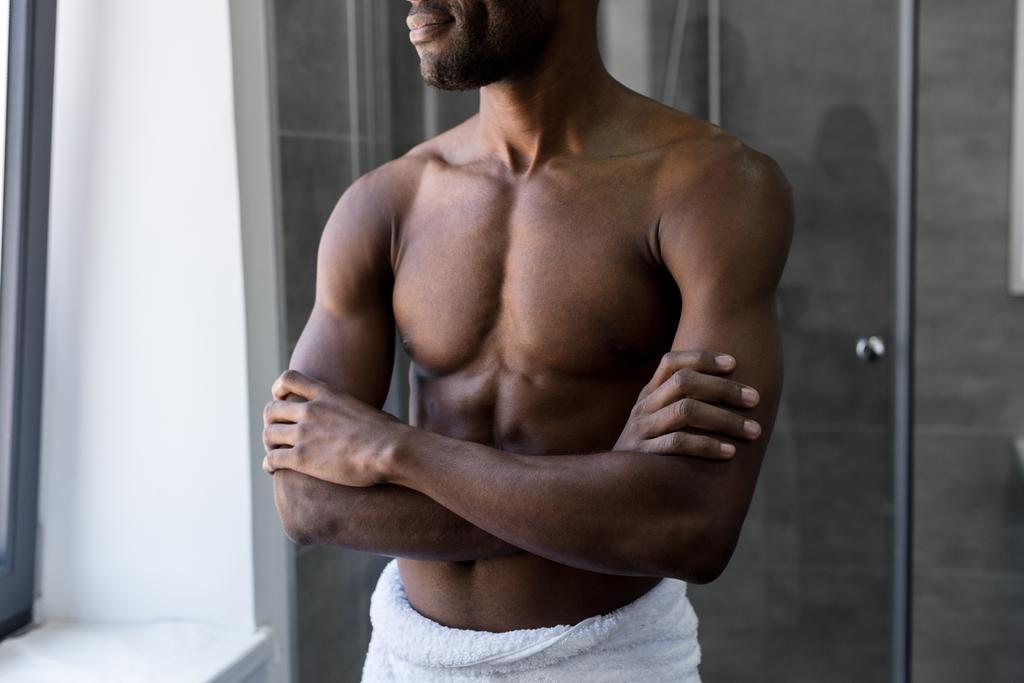 обрізаний знімок усміхненого афроамериканця в рушниці, що стоїть з схрещеними руками у ванній
 - Фото, зображення