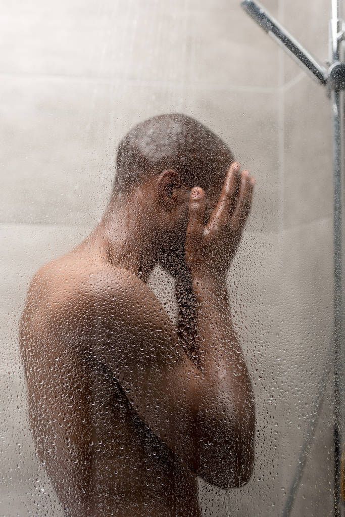 side view of young african american man washing body in shower   - Foto, afbeelding