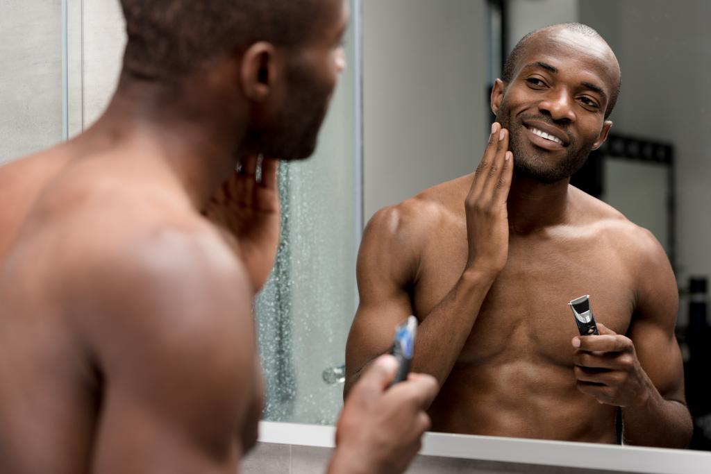sonriente afroamericano hombre afeitándose con trimmer eléctrico en baño
 - Foto, Imagen