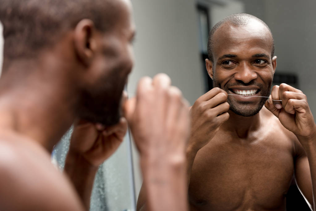 sonriente joven afroamericano hombre sosteniendo hilo dental y mirando al espejo en el baño
 - Foto, Imagen
