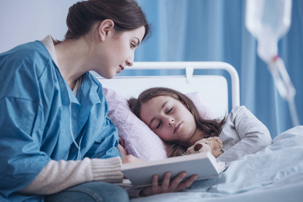 Verzorger lezen van een boek naar een slaperig ziek meisje in de kliniek - Foto, afbeelding