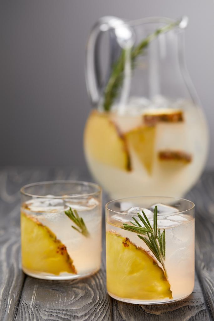 kruik van limonade met ananas en rozemarijn, twee glazen limonade met ananas stukjes, ijsblokjes en rozemarijn op grijs houten tafelblad - Foto, afbeelding