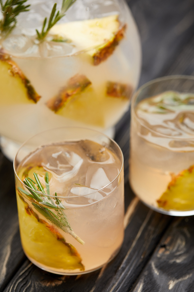 vista parziale della brocca e due bicchieri di limonata con pezzetti di ananas, cubetti di ghiaccio e rosmarino su tavolo grigio in legno
 - Foto, immagini