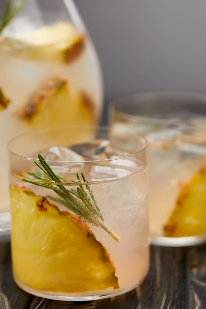 selective focus of jug and two glasses of lemonade with pineapple pieces, ice cubes and rosemary on grey wooden tablet - Photo, Image