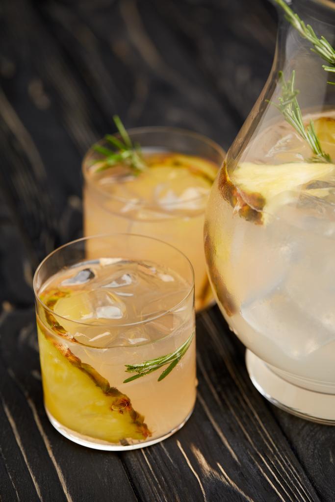 osittainen näkymä kannu ja kaksi lasillista limonadia ananas kappaletta, jääpaloja ja rosmariinia harmaa puinen pöytälevy
 - Valokuva, kuva