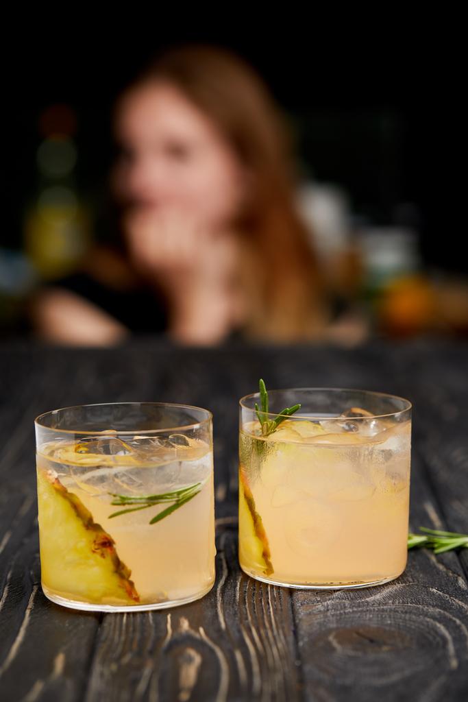 selektiver Fokus von Ananasstücken, Eiswürfeln, Rosmarin in zwei Gläsern Limonade und Frau auf verschwommenem Hintergrund  - Foto, Bild