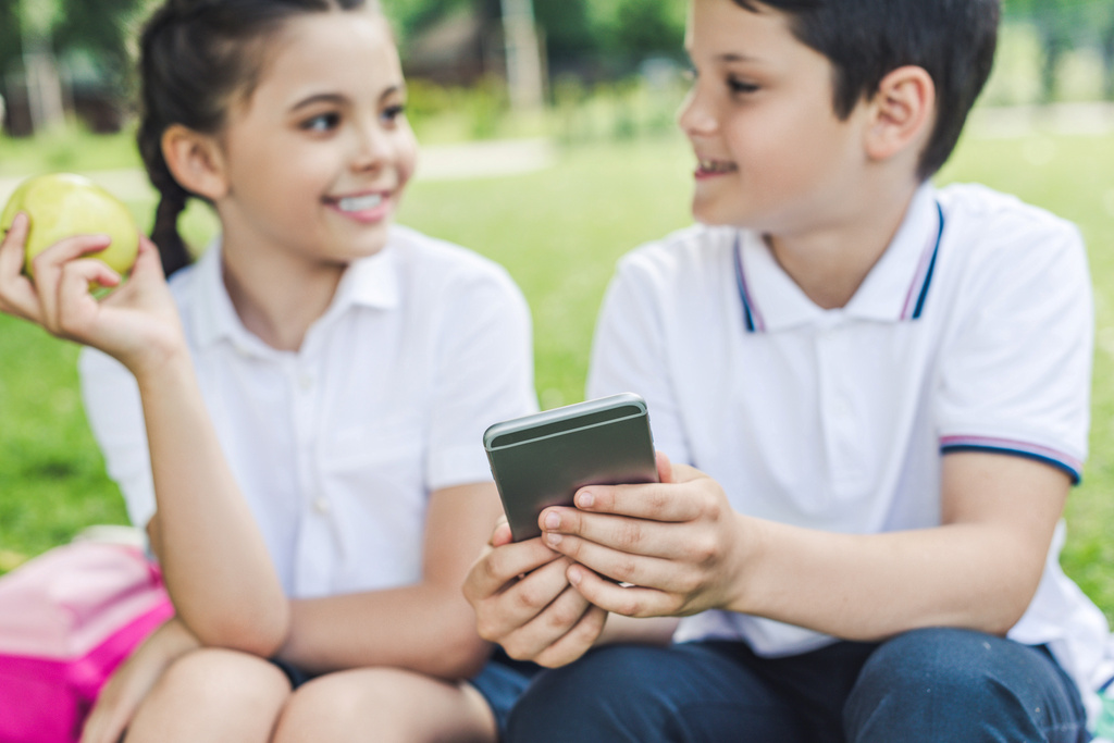 schoolchildren using smartphone together and chatting while sitting on grass - Photo, Image