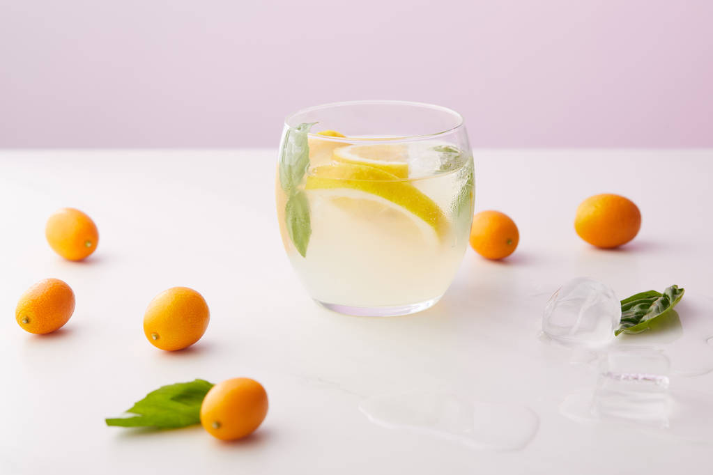 ποτήρι λεμονάδα με φύλλα δυόσμου και φέτες λεμονιού περιβάλλεται από παγάκια, κουμκουάτ σε μοβ φόντο  - Φωτογραφία, εικόνα