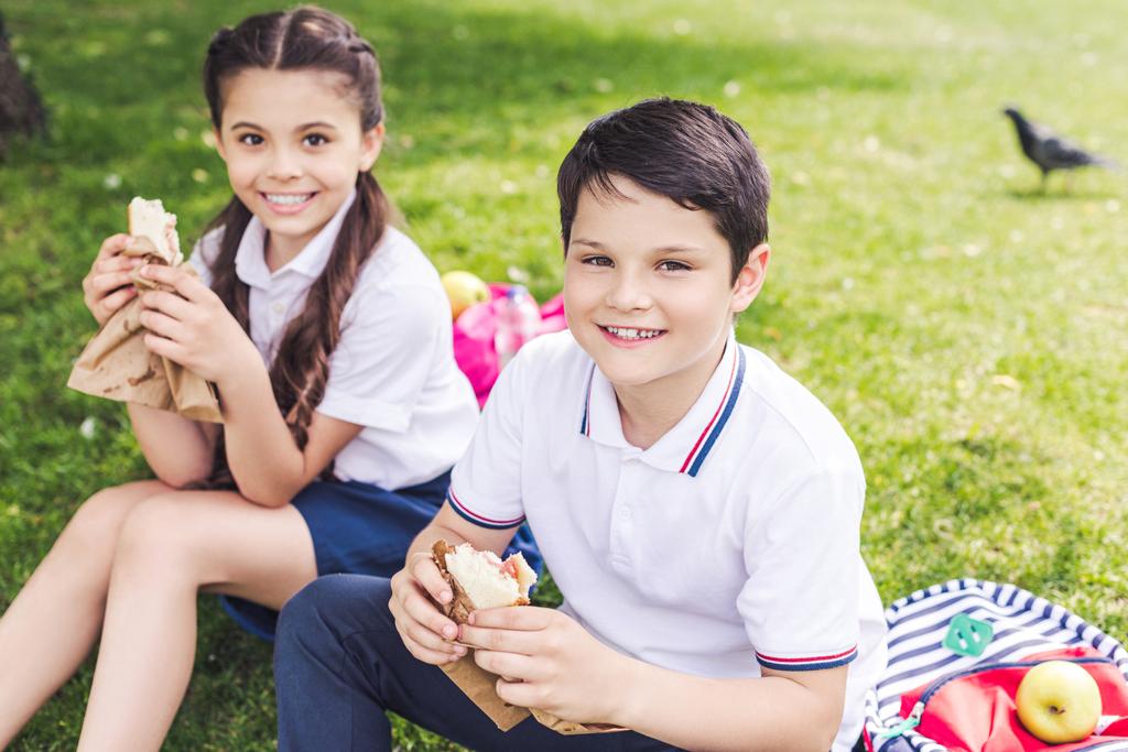 草の上に座って、サンドイッチを食べて笑顔の小学生 - 写真・画像