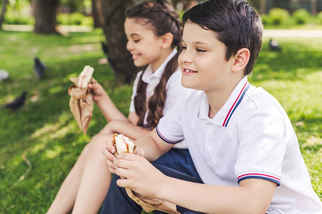 happy schoolchildren sitting on grass and eating sandwiches - Photo, Image