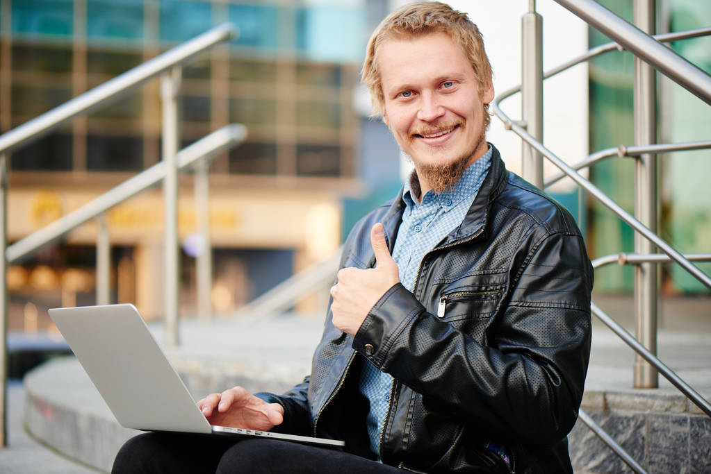 Lächelnder blonder Geschäftsmann in Lederjacke mit Laptop, der Daumen nach oben zeigt, während er auf der Veranda des Business Centers sitzt - Foto, Bild