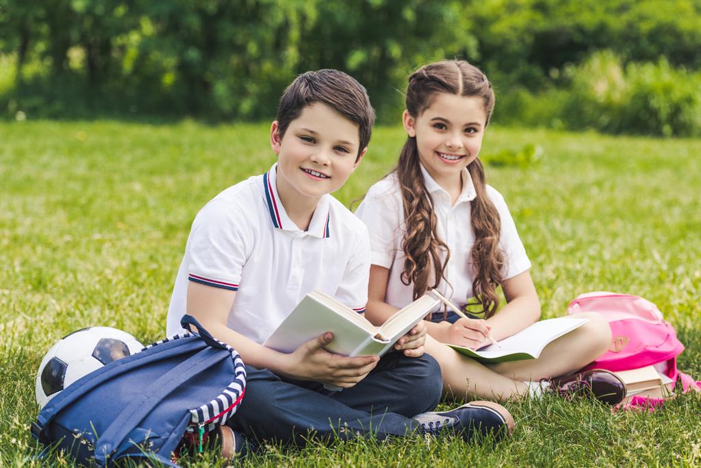 hymyilevät koululaiset tekevät läksyjä yhdessä istuessaan nurmikolla puistossa ja katsoessaan kameraa
 - Valokuva, kuva