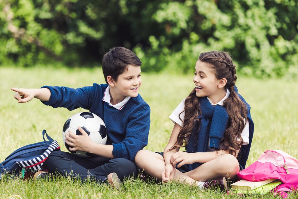 šťastné děti sedí na trávě v parku s batohy a fotbalový míč a chatování - Fotografie, Obrázek