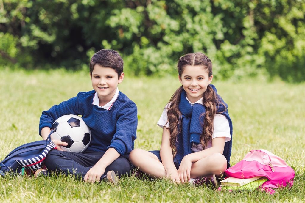 onnellinen koululaiset istuu ruoho puistossa reput ja jalkapallo pallo
 - Valokuva, kuva