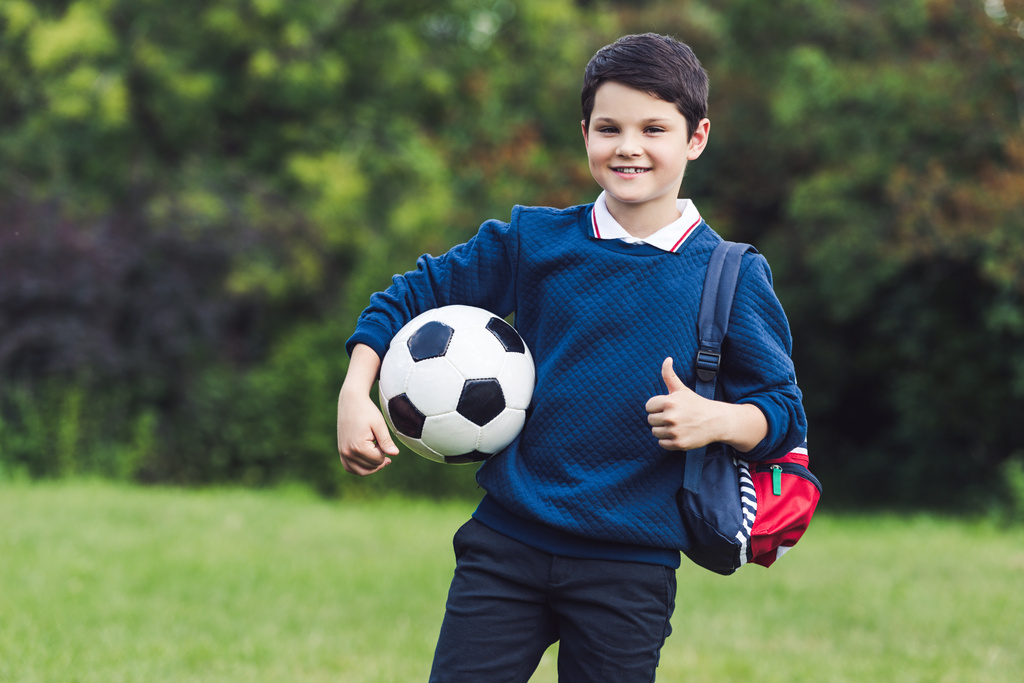 Glückliches Kind mit Fußball und Rucksack zeigt Daumen hoch auf Rasenplatz - Foto, Bild