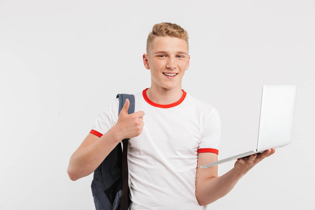 Foto van vrolijke mannelijke student dragen t-shirt en rugzak duim opdagen terwijl opengeklapte laptop geïsoleerd op witte achtergrond - Foto, afbeelding