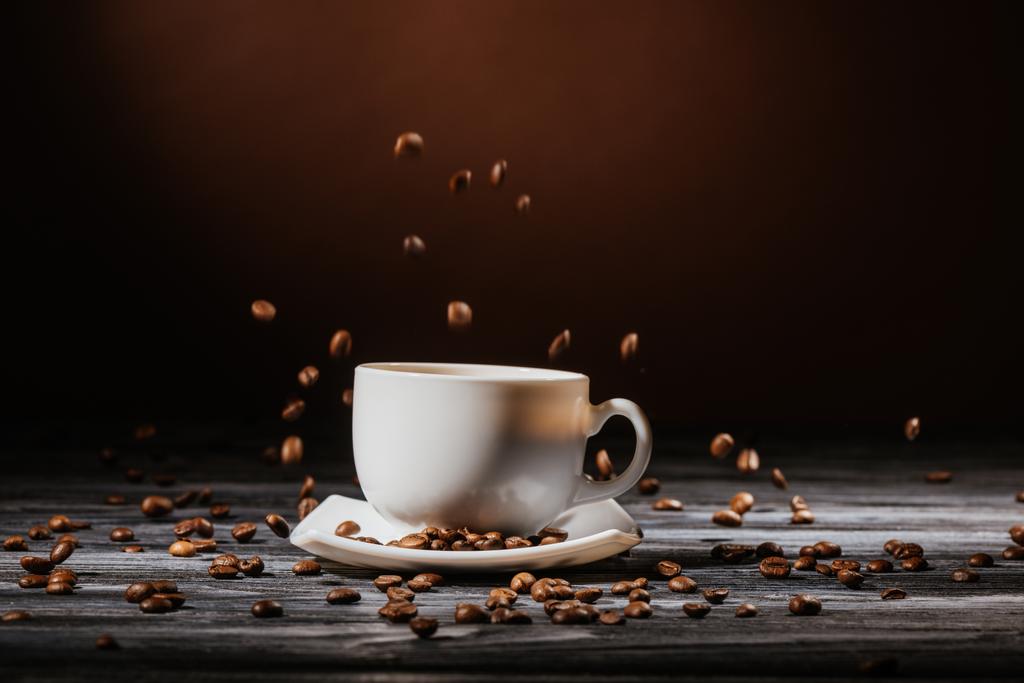 close-up shot of cup with coffee beans falling around on rustic wooden table - Photo, Image