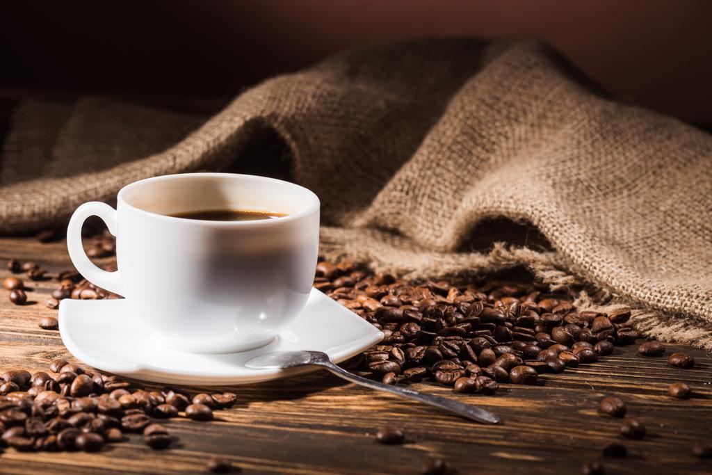 Tasse Kaffee mit gerösteten Bohnen und Sacktuch auf Holztisch - Foto, Bild