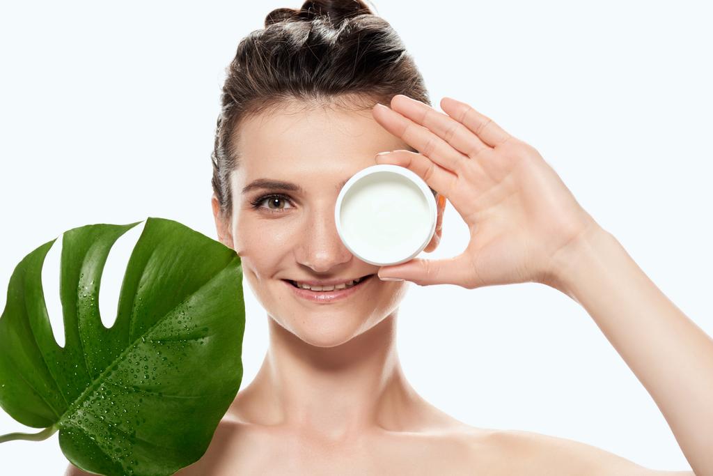 mooie vrouw met gezichtscrème in plastic container en monstera blad, geïsoleerd op wit - Foto, afbeelding